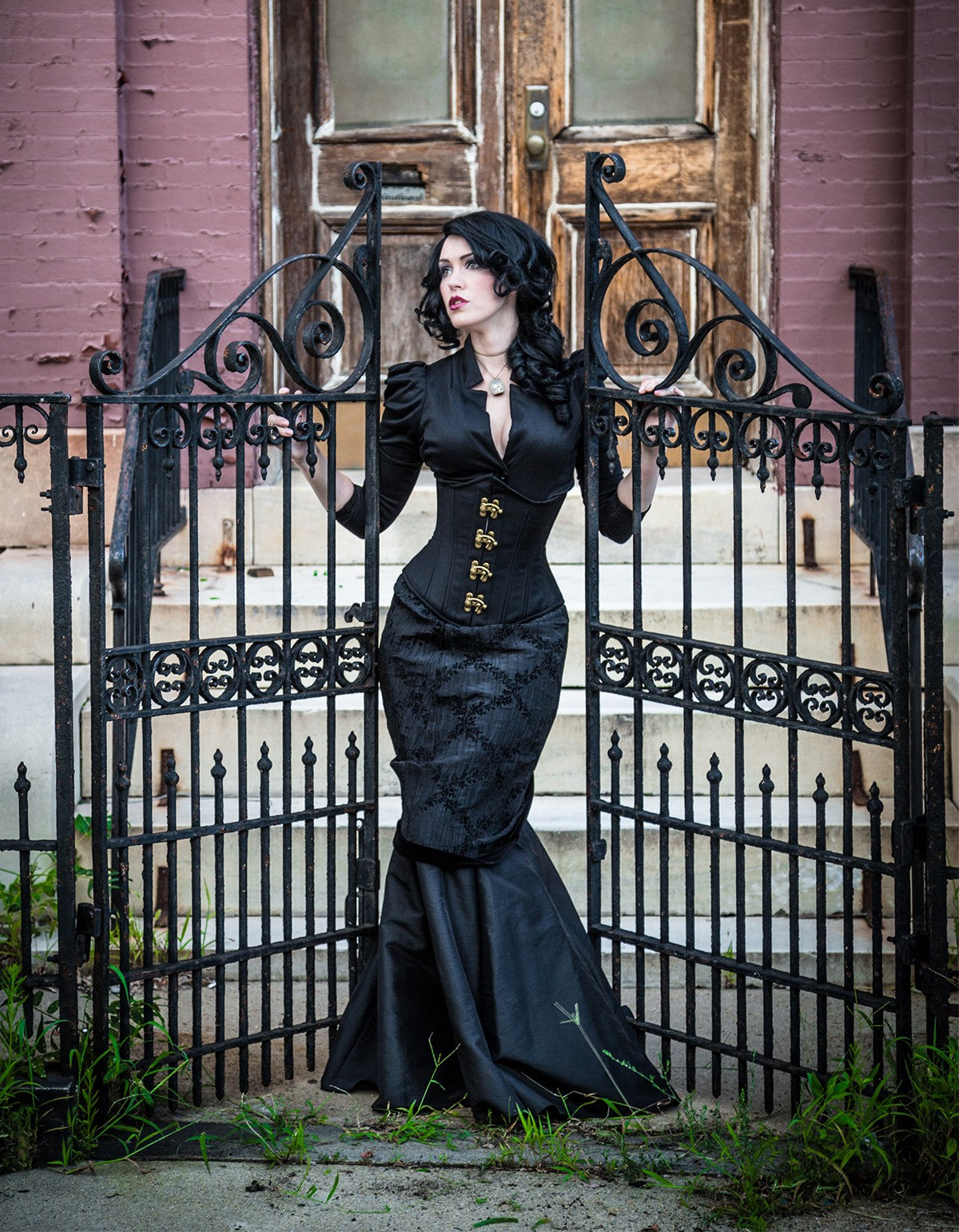Gothic Cashmere Corset, slim silhouette, regular underbust length, front view with brass swing clasps on model standing by wrought iron gates